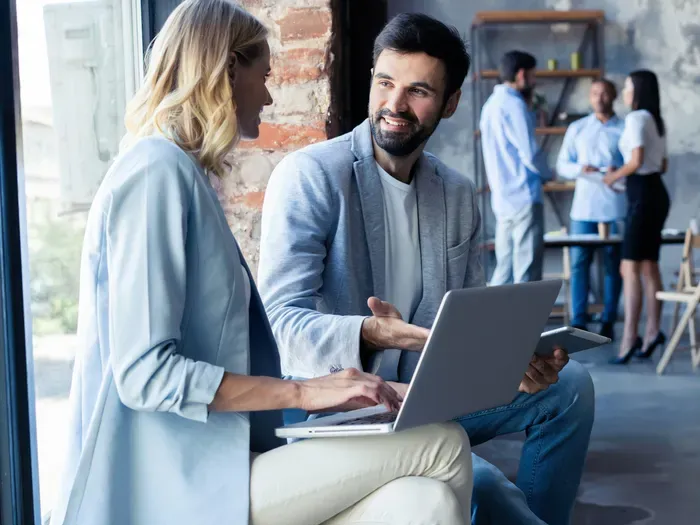 Two professionals having discussion with laptop in modern office with colleagues collaborating in background
